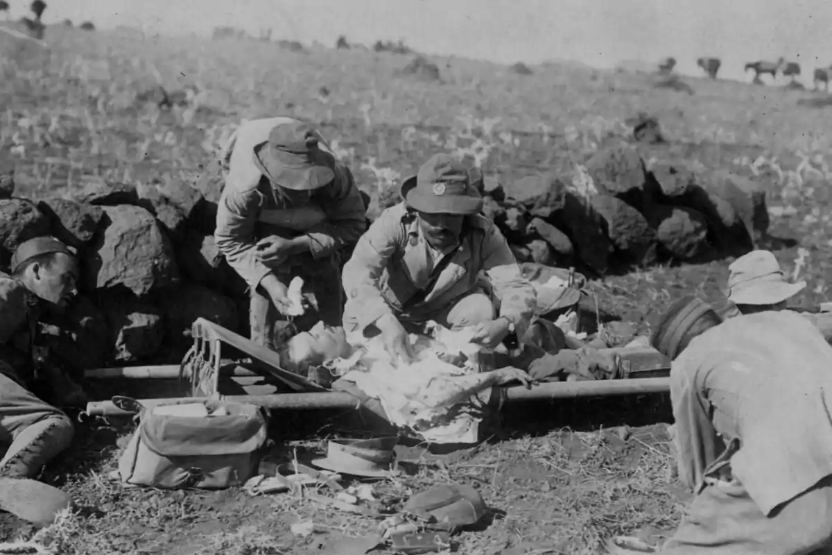 Soldados atienden a un compañero herido en el campo durante una guerra, rodeados de equipo médico improvisado y piedras. Soldados atienden a un compañero herido en el campo durante una guerra, rodeados de equipo médico improvisado y piedras.
