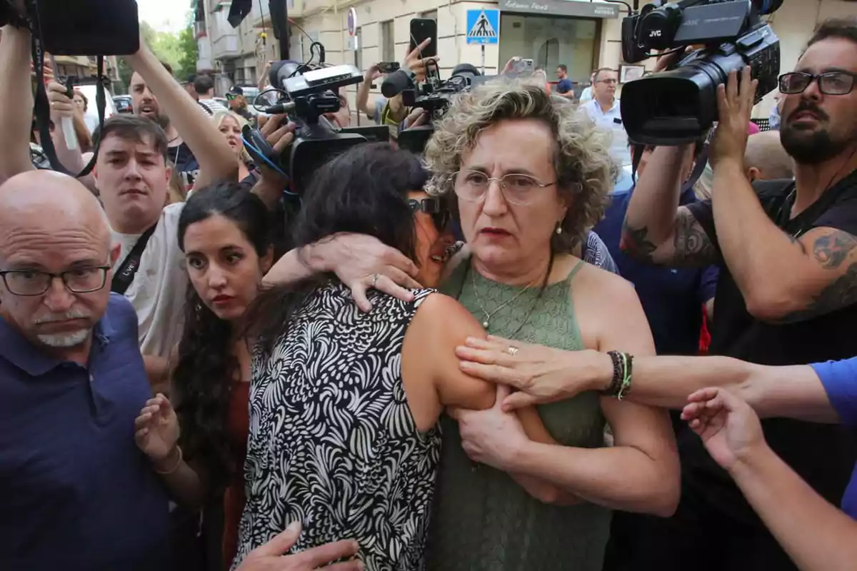 Una mujer abraza a otra con expresión seria mientras varias personas y cámaras de prensa las rodean en la calle Una mujer abraza a otra con expresión seria mientras varias personas y cámaras de prensa las rodean en la calle