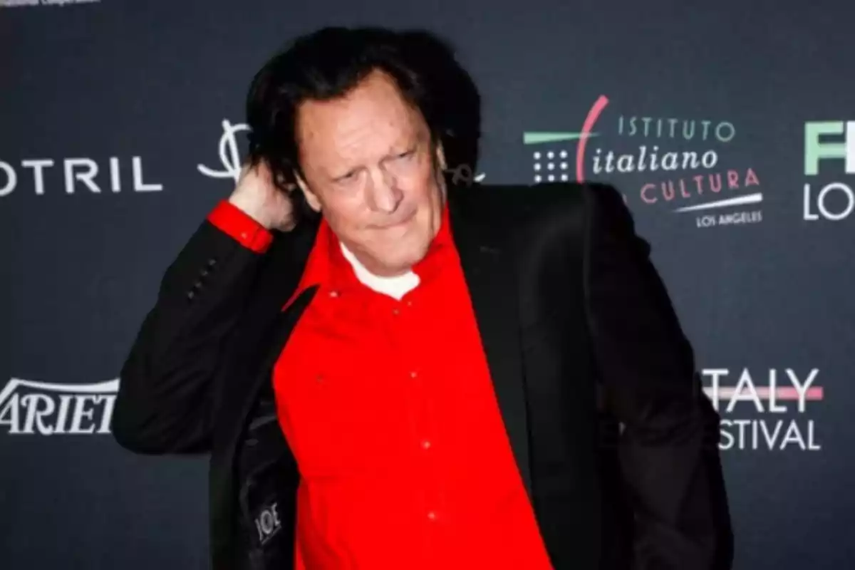 Hombre de cabello oscuro con camisa roja y saco negro posando frente a un fondo con logotipos de un evento