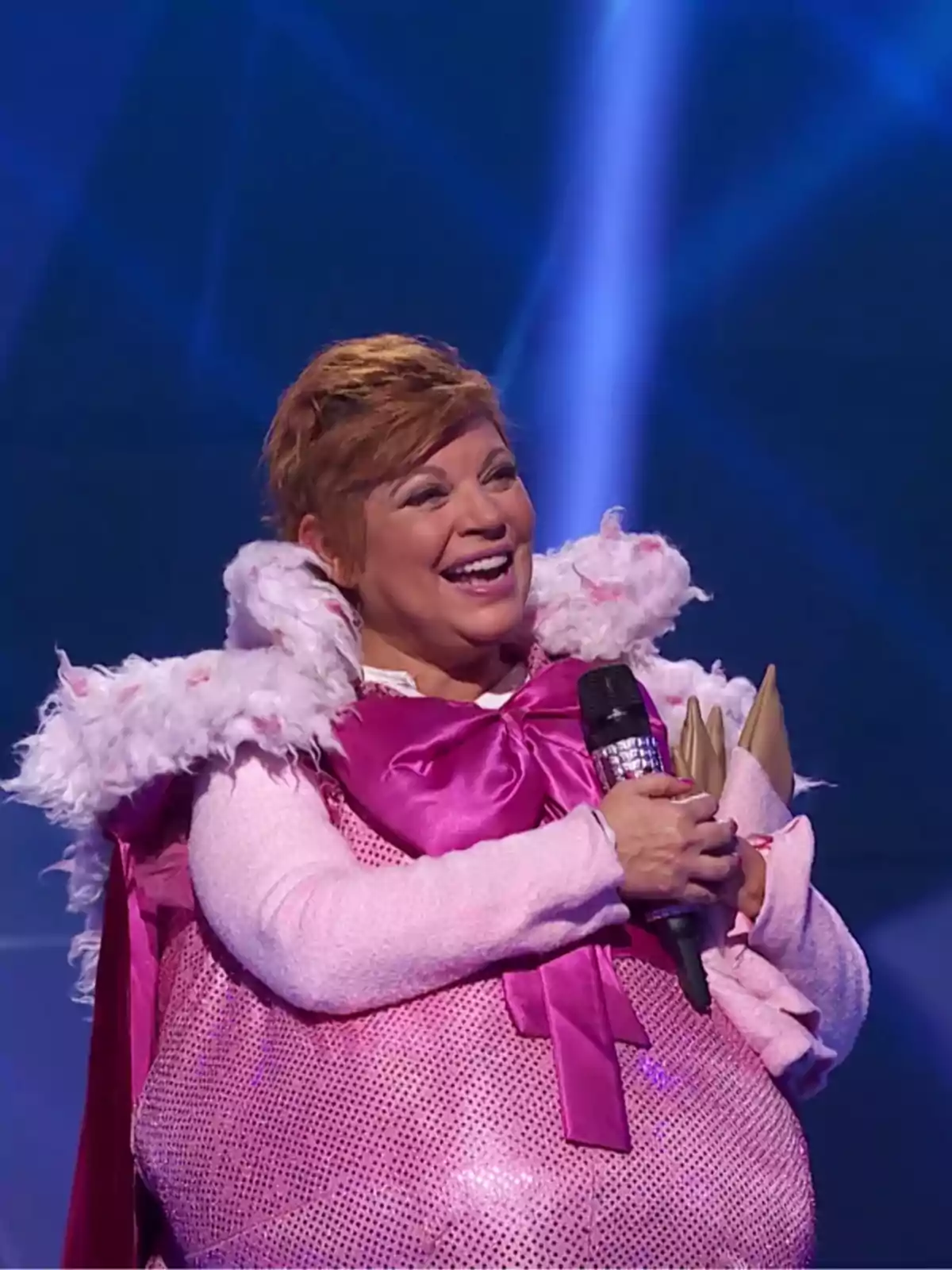 Persona sonriente con disfraz rosa brillante y lazo grande sosteniendo un micrófono en un escenario iluminado Persona sonriente con disfraz rosa brillante y lazo grande sosteniendo un micrófono en un escenario iluminado