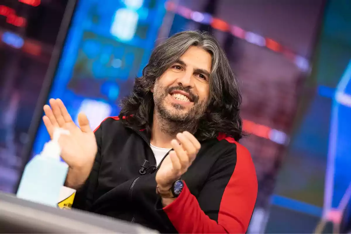 Hombre de cabello largo y barba sonríe mientras aplaude en un estudio de televisión con fondo colorido y desenfocado Hombre de cabello largo y barba sonríe mientras aplaude en un estudio de televisión con fondo colorido y desenfocado