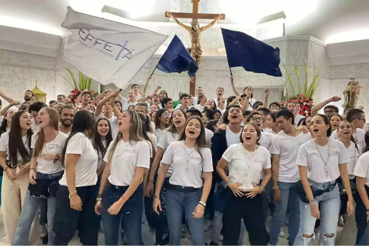 Grupo numeroso de jóvenes con camisetas blancas celebrando y cantando dentro de una iglesia, algunos sostienen banderas y al fondo se observa un crucifijo grande. Grupo numeroso de jóvenes con camisetas blancas celebrando y cantando dentro de una iglesia, algunos sostienen banderas y al fondo se observa un crucifijo grande.