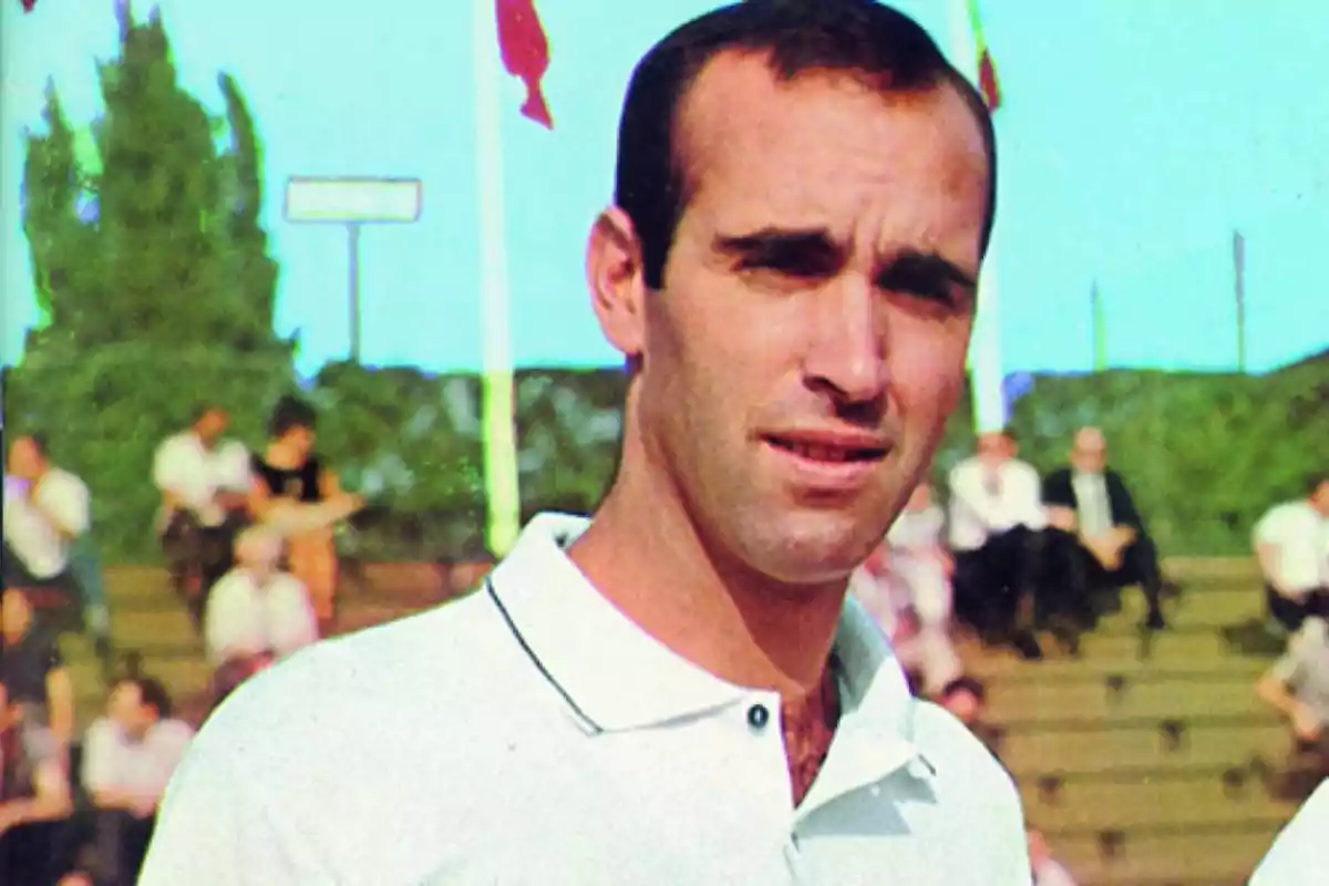 Hombre de cabello corto y oscuro con camisa blanca posando al aire libre frente a gradas con personas al fondo Hombre de cabello corto y oscuro con camisa blanca posando al aire libre frente a gradas con personas al fondo