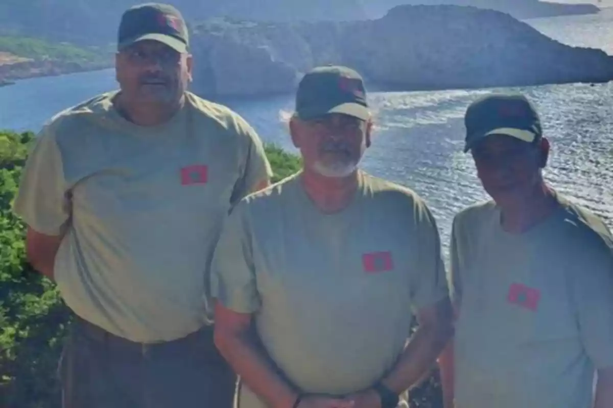 Tres hombres con camisetas y gorras verdes posan juntos al aire libre con un paisaje de mar y montañas al fondo