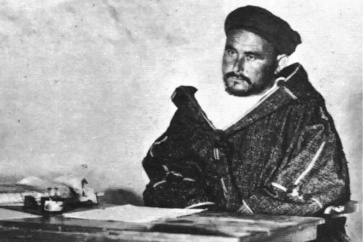 Hombre con barba y boina sentado en un escritorio con papeles y una taza, vistiendo ropa tradicional