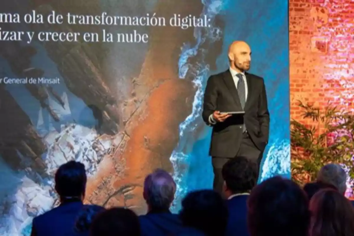 Un hombre con traje sostiene una tableta mientras da una presentación frente a una audiencia, con una pantalla grande de fondo que muestra un mensaje sobre transformación digital y la nube Un hombre con traje sostiene una tableta mientras da una presentación frente a una audiencia, con una pantalla grande de fondo que muestra un mensaje sobre transformación digital y la nube