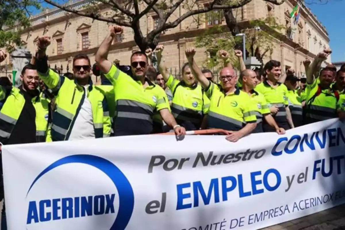 Trabajadores con chalecos amarillos y grises levantan el puño mientras sostienen una pancarta de Acerinox en una manifestación en la calle frente a un edificio antiguo Trabajadores con chalecos amarillos y grises levantan el puño mientras sostienen una pancarta de Acerinox en una manifestación en la calle frente a un edificio antiguo
