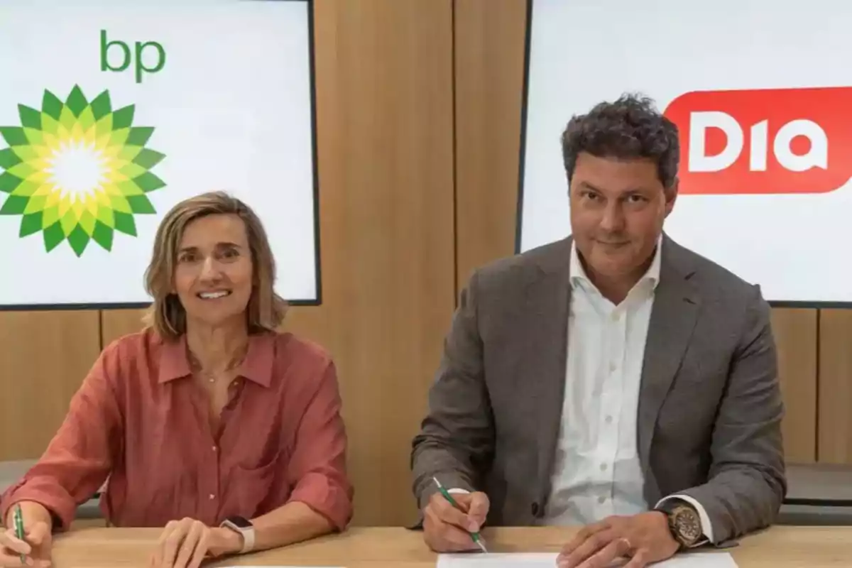 Dos personas sentadas en una mesa firmando documentos con los logotipos de BP y Dia en pantallas al fondo