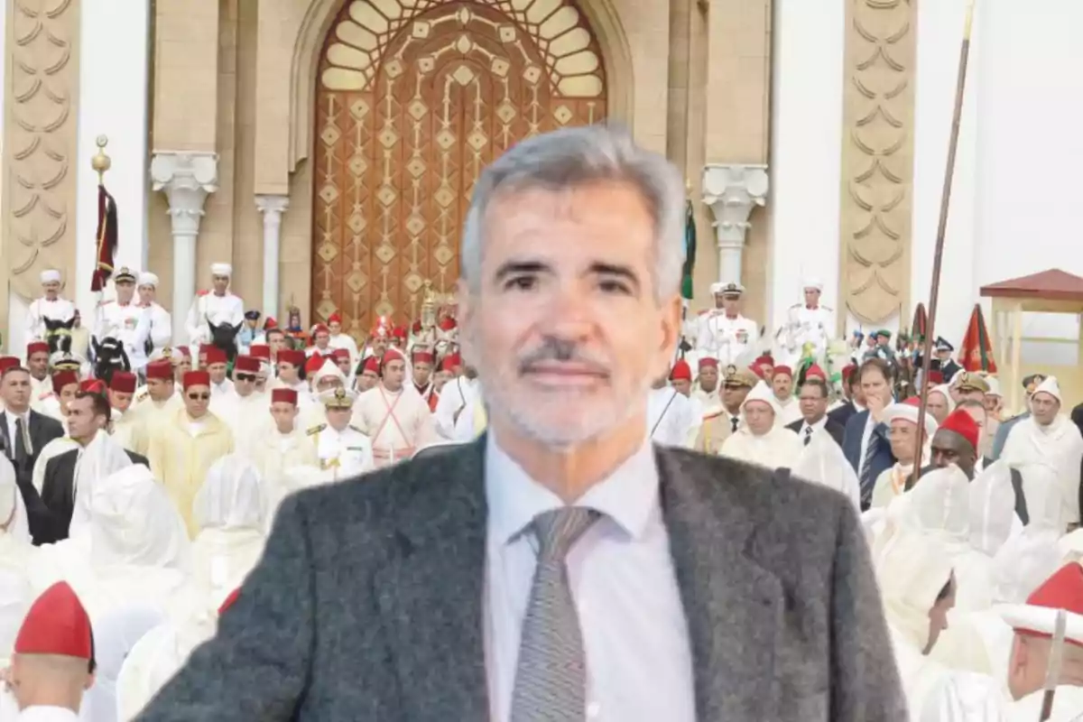 Hombre de cabello canoso y barba con traje gris y corbata posando frente a una multitud de personas vestidas con trajes tradicionales y militares en un evento solemne frente a una puerta ornamentada