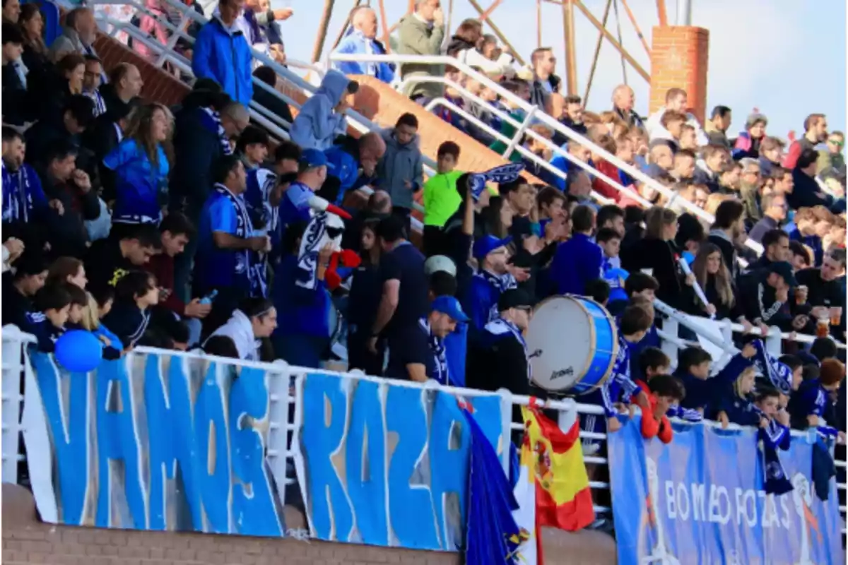 Aficionados animando en las gradas de un estadio con pancartas azules, banderas y un tambor Aficionados animando en las gradas de un estadio con pancartas azules, banderas y un tambor