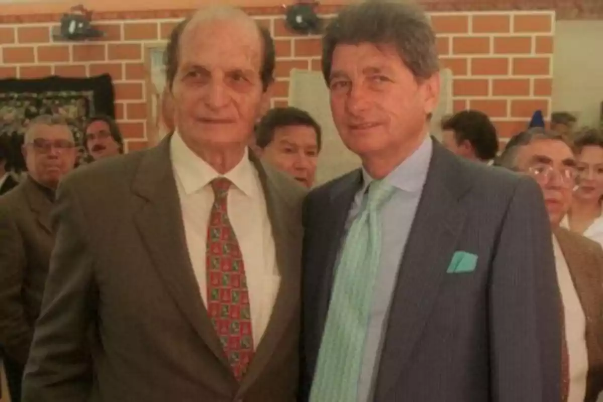 Dos hombres de traje posan juntos en un evento social con varias personas al fondo Dos hombres de traje posan juntos en un evento social con varias personas al fondo