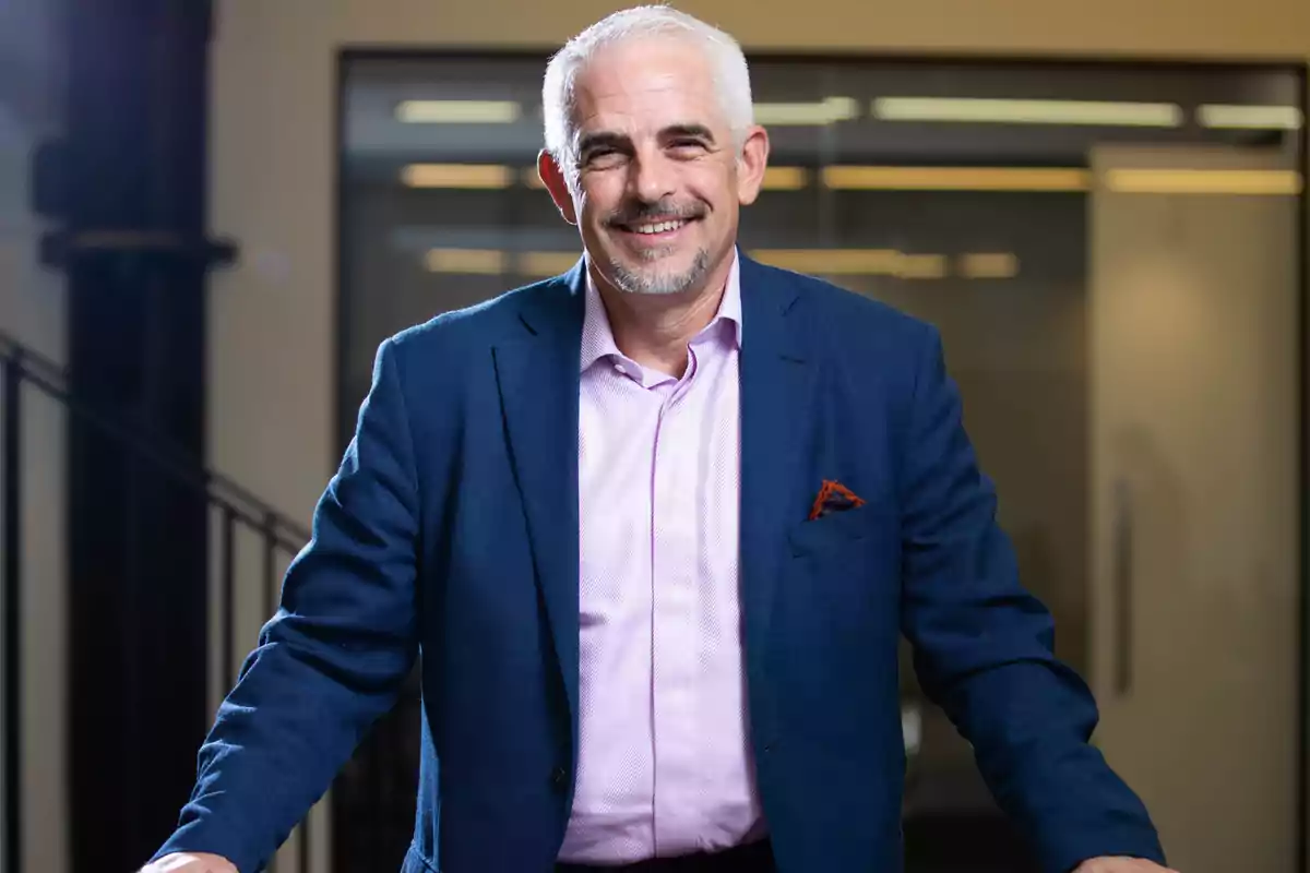 Hombre de cabello canoso y barba corta, vestido con saco azul y camisa rosa, sonriendo en una oficina moderna Hombre de cabello canoso y barba corta, vestido con saco azul y camisa rosa, sonriendo en una oficina moderna