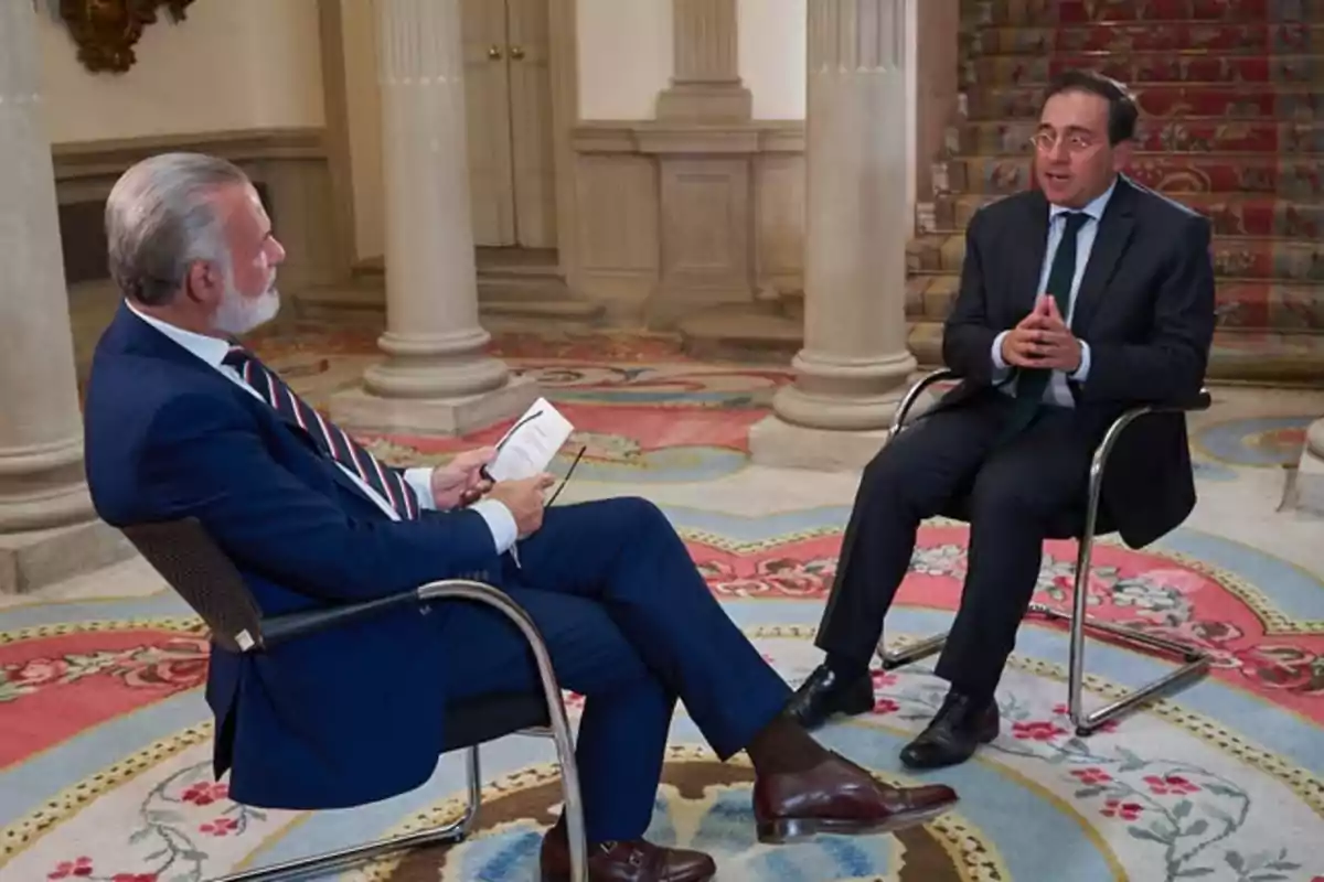 Dos hombres con traje conversan sentados en sillas dentro de un elegante salón con alfombra colorida y columnas al fondo Dos hombres con traje conversan sentados en sillas dentro de un elegante salón con alfombra colorida y columnas al fondo