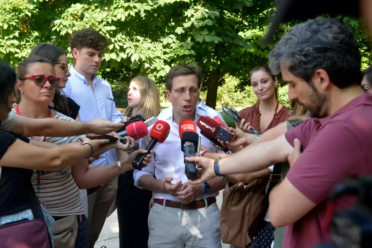 Un hombre rodeado de periodistas sostiene una rueda de prensa al aire libre mientras varias personas le acercan micrófonos y grabadoras