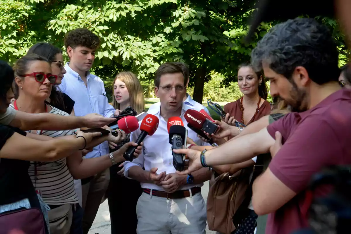Un hombre rodeado de periodistas sostiene una rueda de prensa al aire libre mientras varios micrófonos se acercan hacia él
