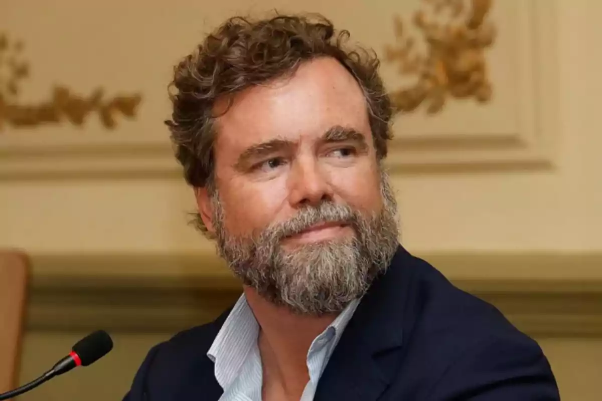 Hombre de barba y cabello rizado con saco azul sentado frente a un micrófono en una sala decorada elegantemente