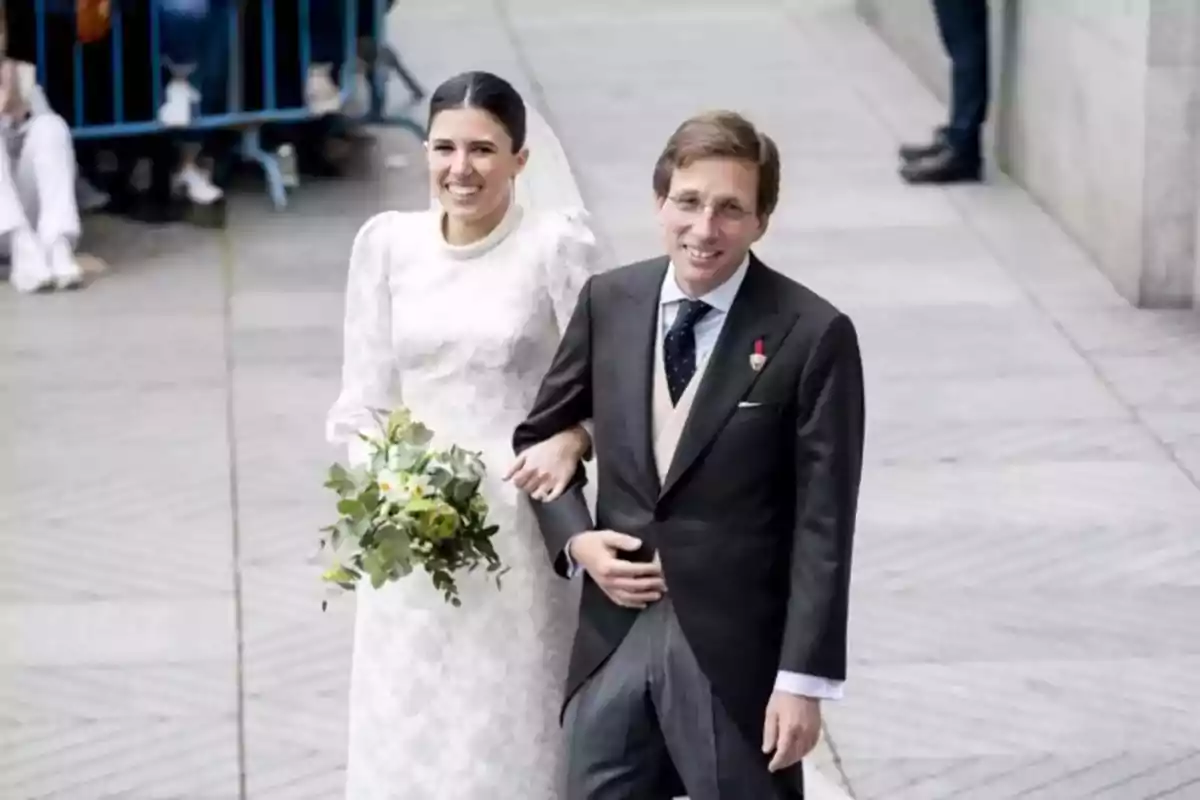 Pareja sonriente vestida de novios caminando juntos al aire libre mientras la novia sostiene un ramo de flores