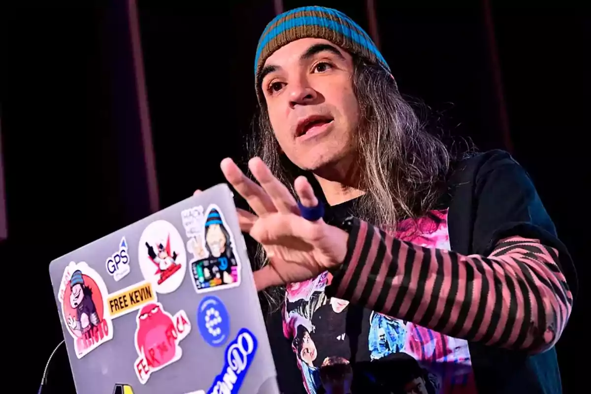 Persona con cabello largo y gorro de rayas hablando mientras usa una laptop decorada con varias calcomanías coloridas Persona con cabello largo y gorro de rayas hablando mientras usa una laptop decorada con varias calcomanías coloridas