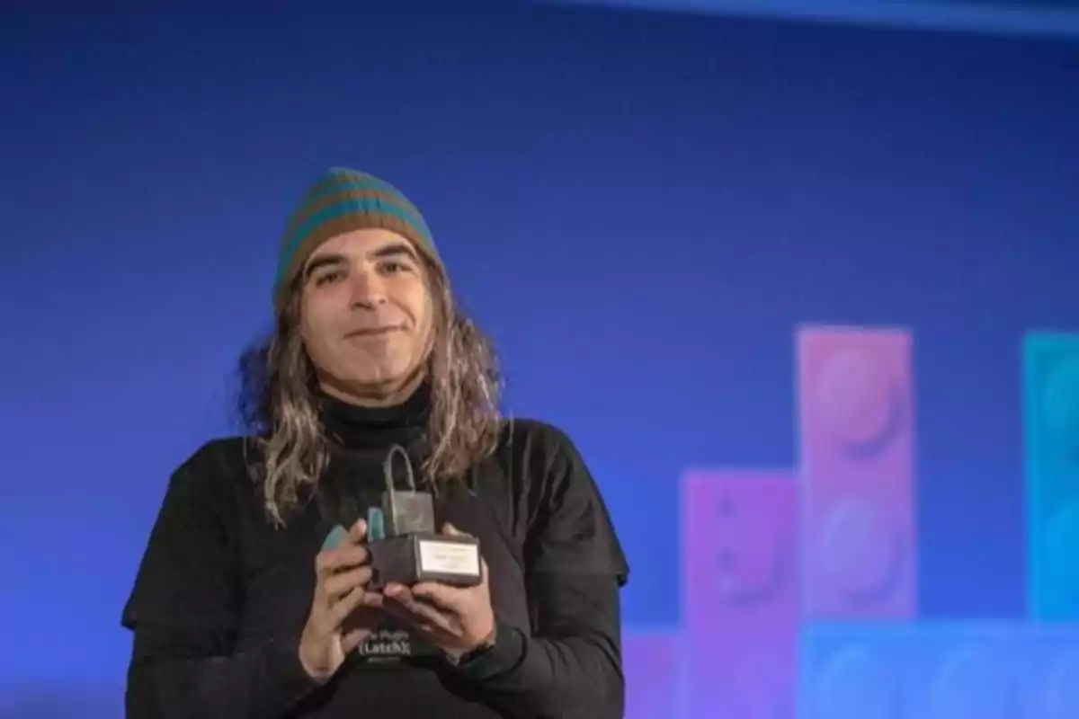 Persona con gorro de rayas y cabello largo sostiene un trofeo en un escenario con fondo azul y figuras geométricas de colores. Persona con gorro de rayas y cabello largo sostiene un trofeo en un escenario con fondo azul y figuras geométricas de colores.