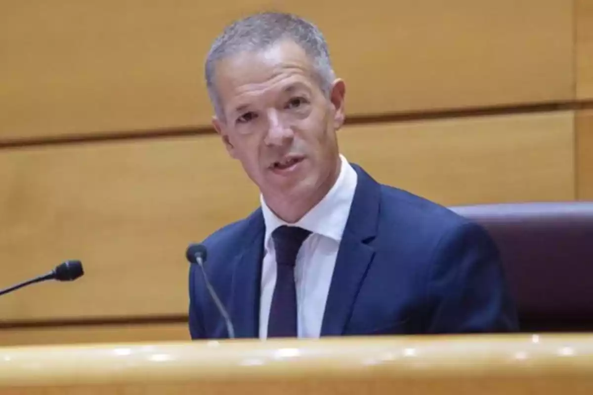 Hombre de cabello corto y canoso con traje azul y corbata hablando frente a un micrófono en un entorno institucional Hombre de cabello corto y canoso con traje azul y corbata hablando frente a un micrófono en un entorno institucional