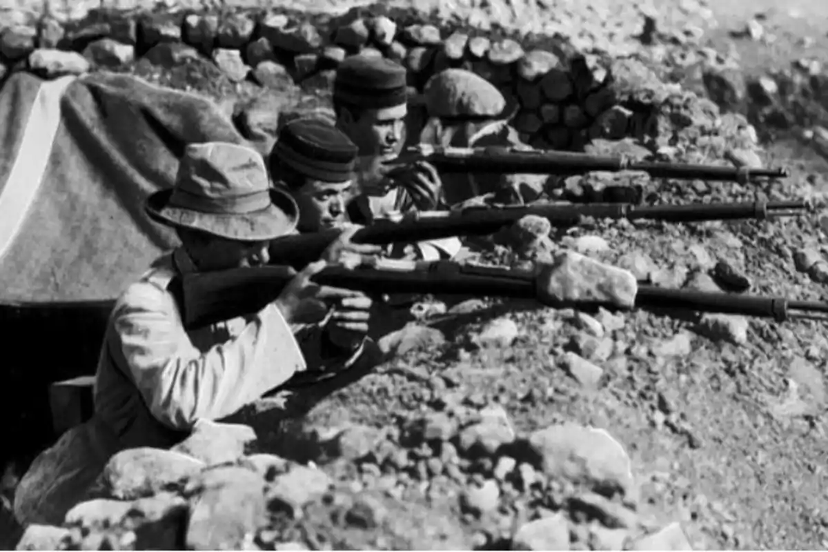 Cuatro soldados apuntan con rifles desde una trinchera de piedra en un entorno árido
