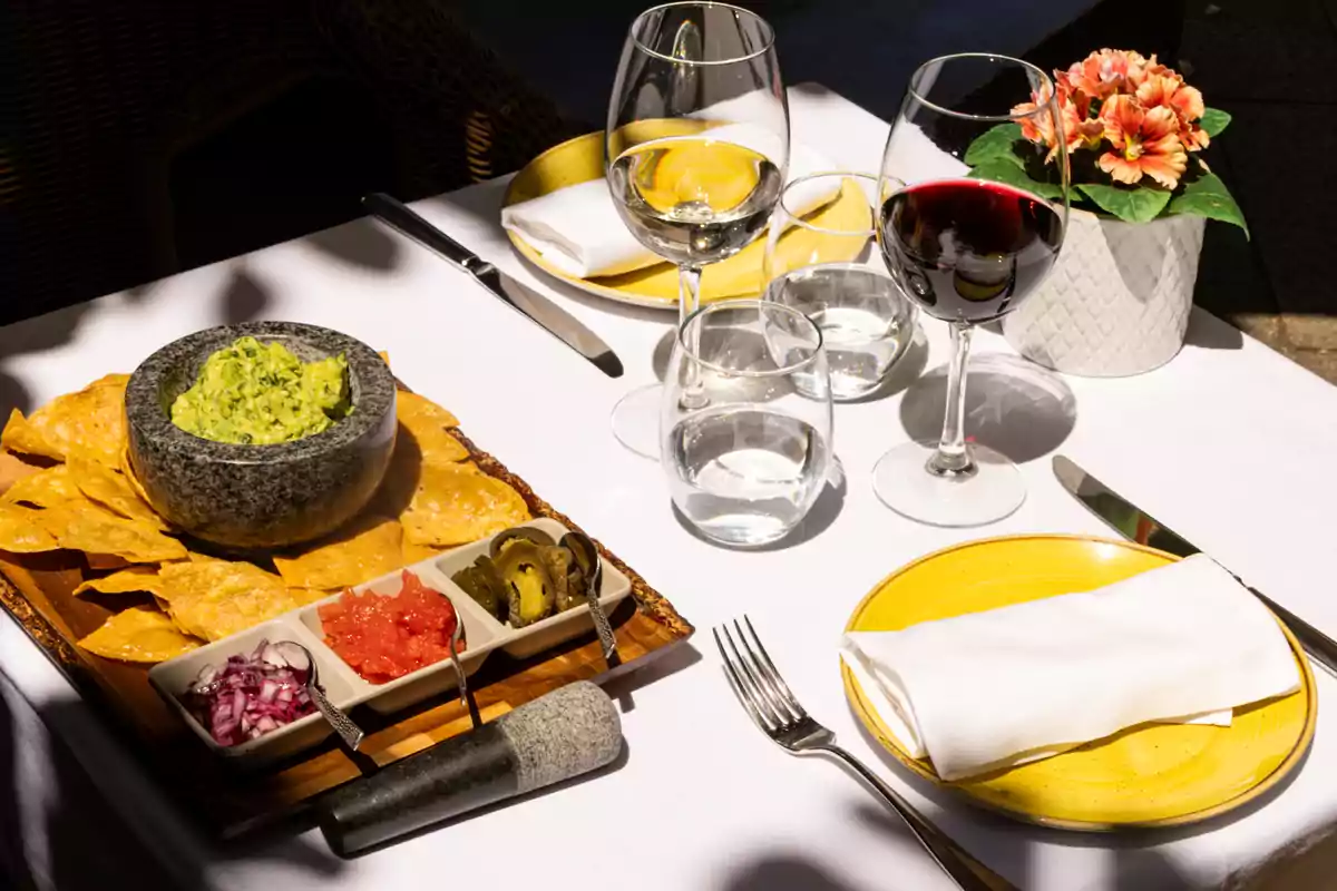 Mesa elegante con guacamole en molcajete, totopos, salsas, copas de vino tinto y agua, platos amarillos y una maceta con flores Mesa elegante con guacamole en molcajete, totopos, salsas, copas de vino tinto y agua, platos amarillos y una maceta con flores