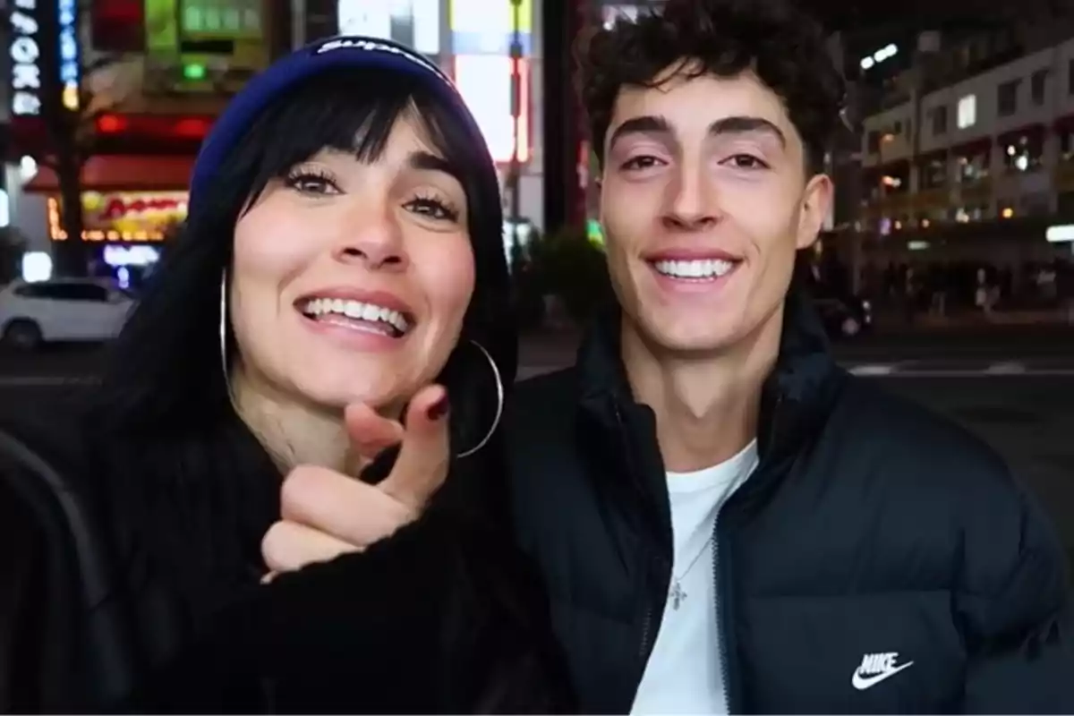 Dos personas jóvenes sonríen mientras posan para una selfie en una ciudad iluminada por luces de neón durante la noche