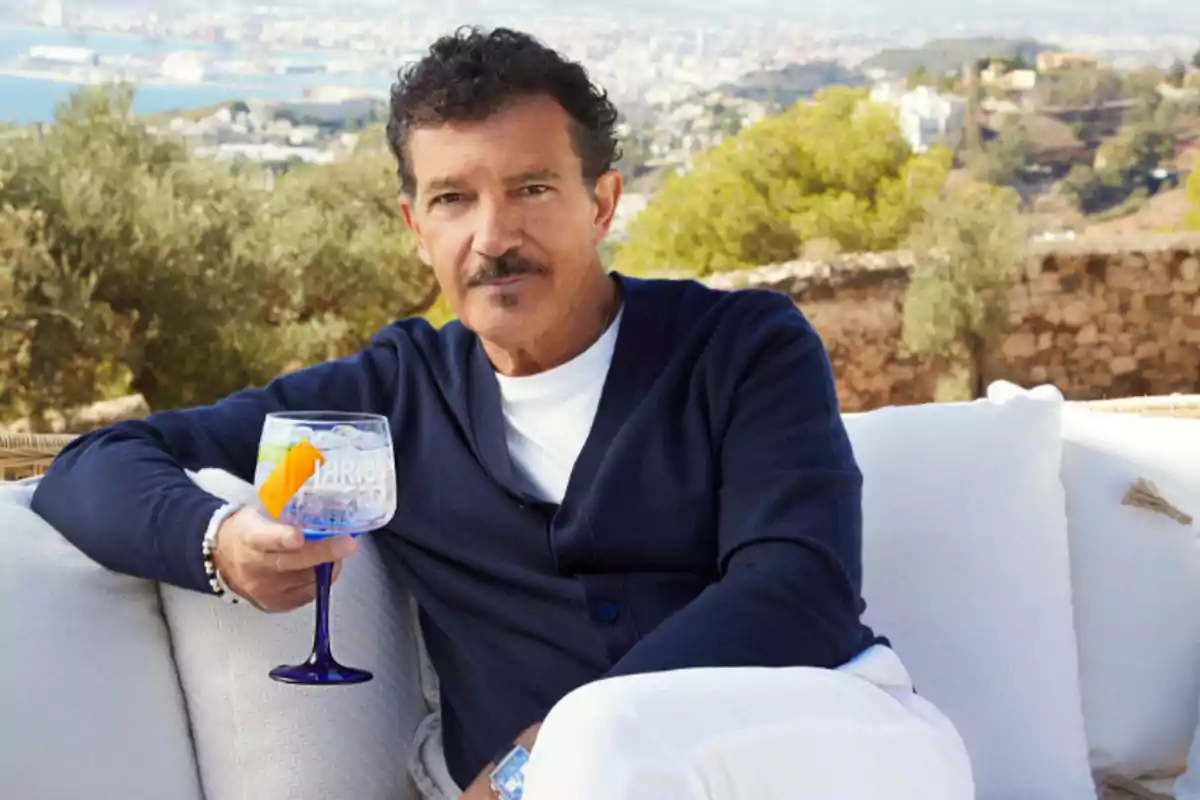 Hombre sentado en un sofá al aire libre sosteniendo una copa de cóctel con hielo y una rodaja de naranja, con un paisaje de ciudad y naturaleza al fondo