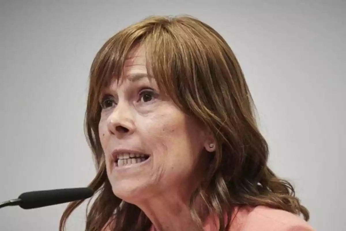 Mujer de cabello castaño hablando frente a un micrófono con expresión seria Mujer de cabello castaño hablando frente a un micrófono con expresión seria