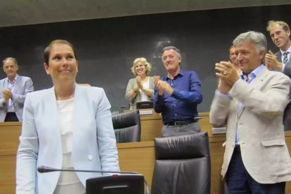 Una mujer de pie en una sala de reuniones mientras varias personas la aplauden y sonríen en el fondo Una mujer de pie en una sala de reuniones mientras varias personas la aplauden y sonríen en el fondo