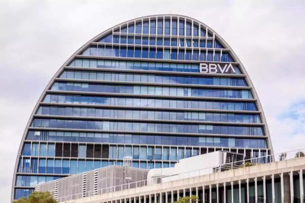 Edificio moderno de oficinas con forma semicircular y fachada de vidrio donde se observa el logo de BBVA en la parte superior Edificio moderno de oficinas con forma semicircular y fachada de vidrio donde se observa el logo de BBVA en la parte superior