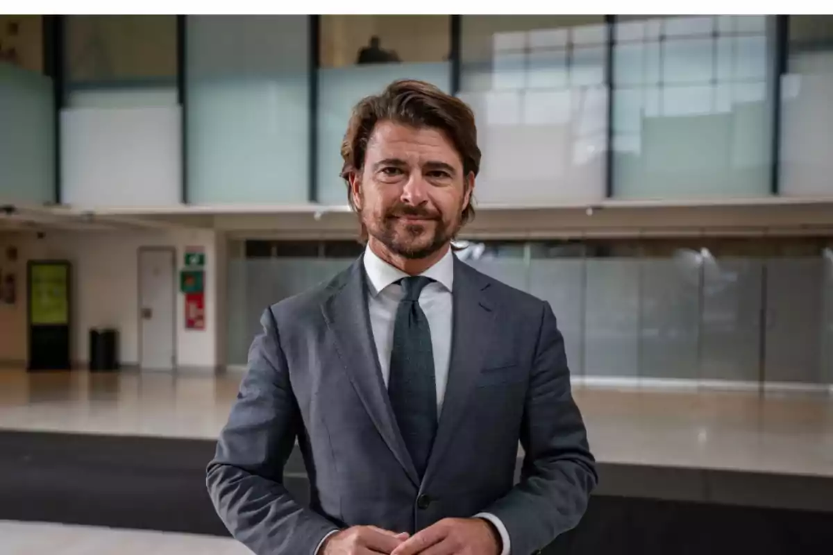 Hombre de cabello castaño y barba vestido con traje gris y corbata posando en el interior de un edificio moderno con paredes de vidrio Hombre de cabello castaño y barba vestido con traje gris y corbata posando en el interior de un edificio moderno con paredes de vidrio