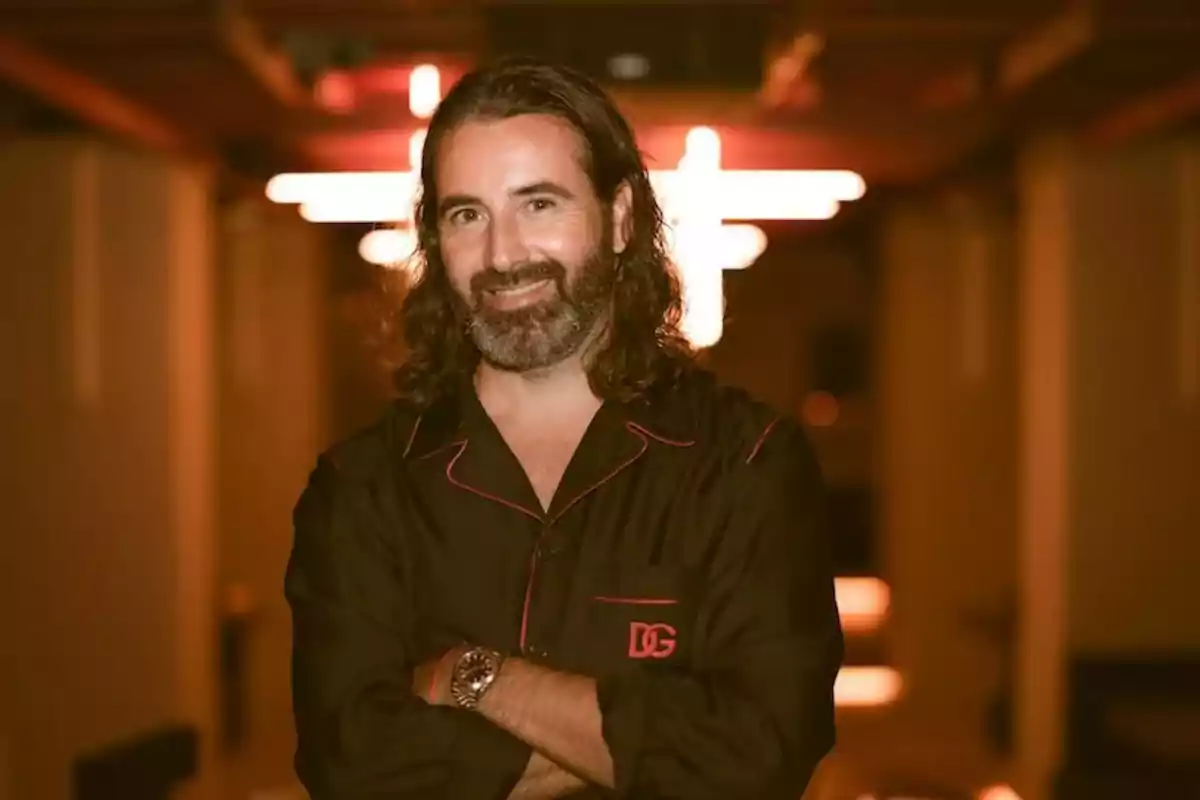 Hombre de cabello largo y barba con camisa oscura sonríe con los brazos cruzados en un ambiente iluminado con luces cálidas Hombre de cabello largo y barba con camisa oscura sonríe con los brazos cruzados en un ambiente iluminado con luces cálidas