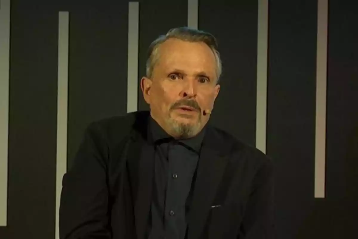 Hombre de mediana edad con barba y bigote usando un micrófono de diadema y vestido con saco oscuro frente a un fondo con líneas verticales claras Hombre de mediana edad con barba y bigote usando un micrófono de diadema y vestido con saco oscuro frente a un fondo con líneas verticales claras