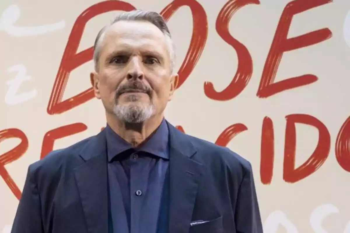 Hombre de cabello canoso y barba corta vestido con saco azul oscuro y camisa azul posando frente a un fondo con letras rojas grandes