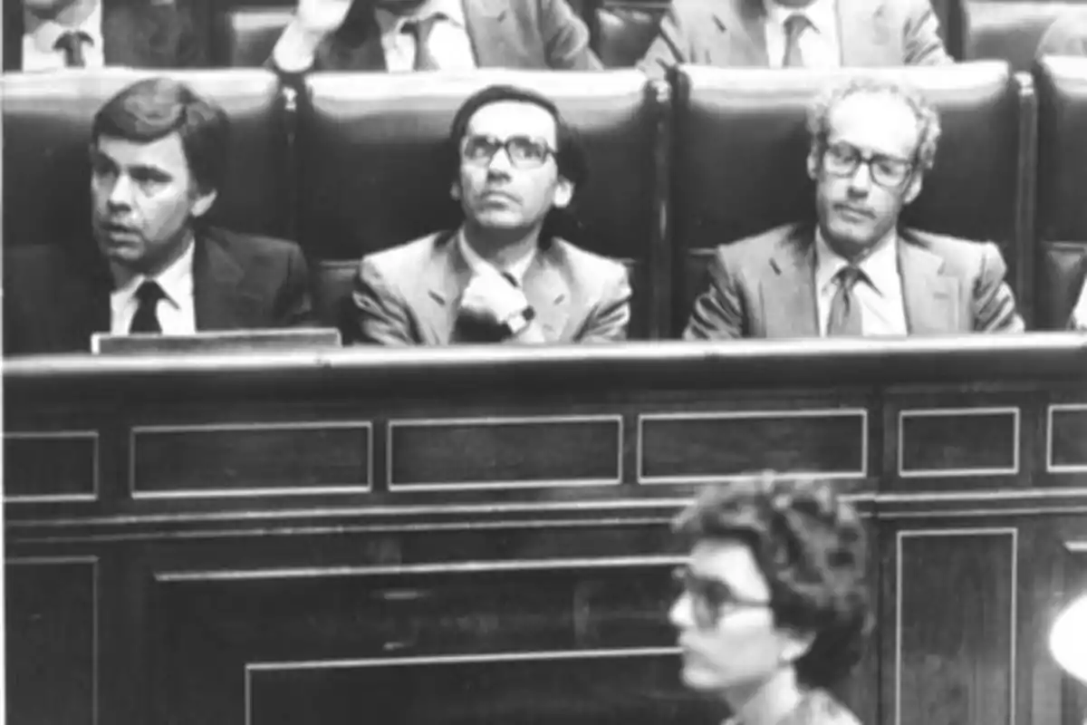 Tres hombres con traje y corbata sentados en un banco de madera en lo que parece ser un parlamento mientras una mujer pasa por delante desenfocada Tres hombres con traje y corbata sentados en un banco de madera en lo que parece ser un parlamento mientras una mujer pasa por delante desenfocada