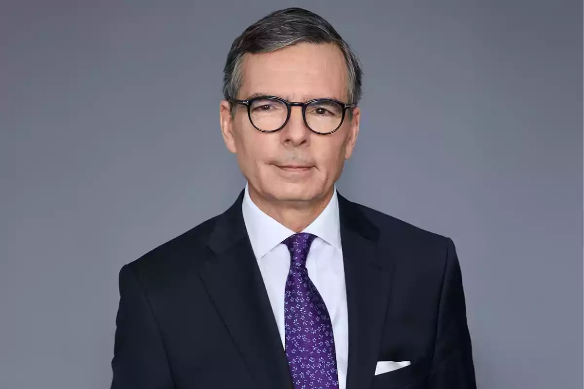 Hombre de mediana edad con gafas y cabello canoso usando traje oscuro, camisa blanca y corbata morada con fondo gris Hombre de mediana edad con gafas y cabello canoso usando traje oscuro, camisa blanca y corbata morada con fondo gris