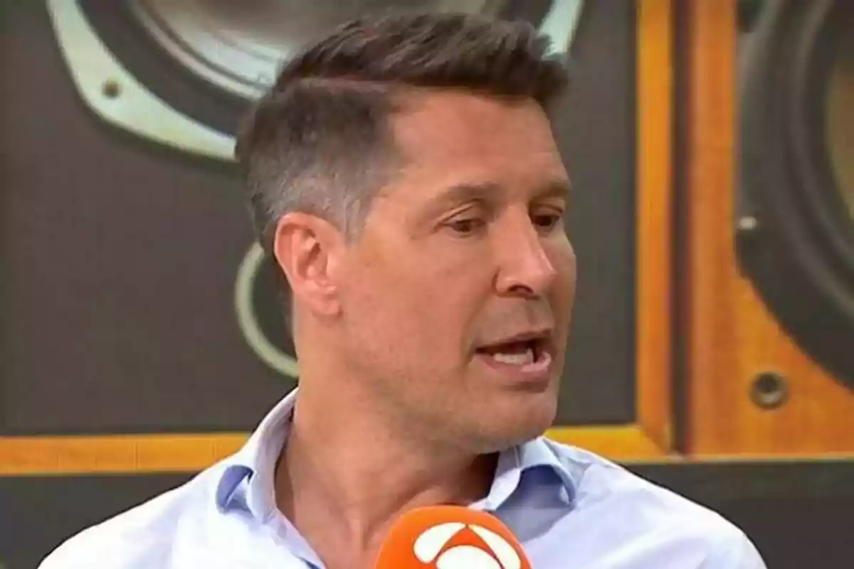 Hombre de cabello corto y canoso hablando frente a un micrófono naranja con fondo de bocinas grandes Hombre de cabello corto y canoso hablando frente a un micrófono naranja con fondo de bocinas grandes