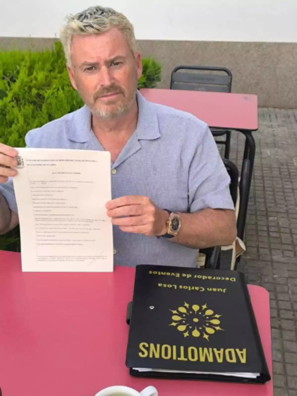 Hombre de cabello rubio y barba sentado en una mesa rosa al aire libre mostrando un documento mientras sostiene una carpeta negra con letras amarillas Hombre de cabello rubio y barba sentado en una mesa rosa al aire libre mostrando un documento mientras sostiene una carpeta negra con letras amarillas