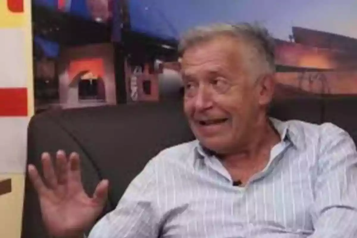 Hombre mayor con camisa clara sentado en un sillón oscuro levantando la mano y sonriendo mientras conversa en un ambiente interior Hombre mayor con camisa clara sentado en un sillón oscuro levantando la mano y sonriendo mientras conversa en un ambiente interior