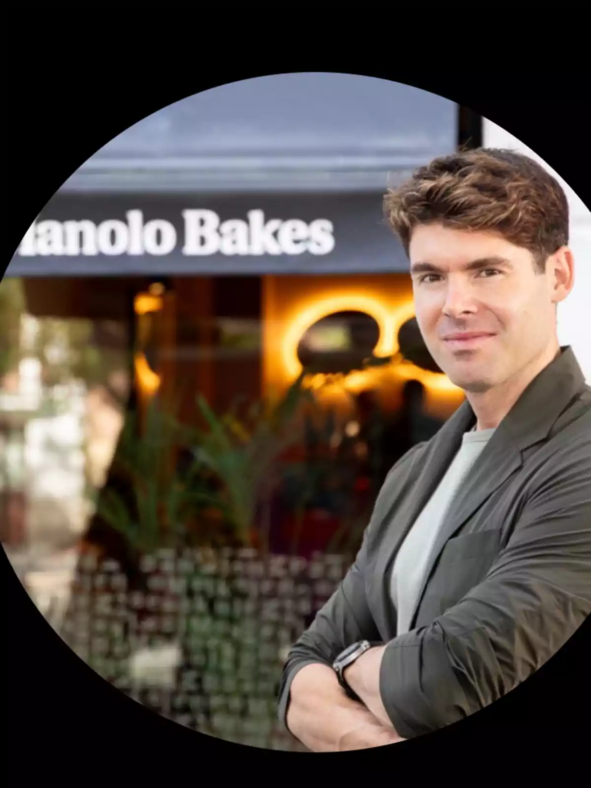 Hombre de cabello castaño con chaqueta gris claro posando frente a una cafetería llamada Manolo Bakes Hombre de cabello castaño con chaqueta gris claro posando frente a una cafetería llamada Manolo Bakes