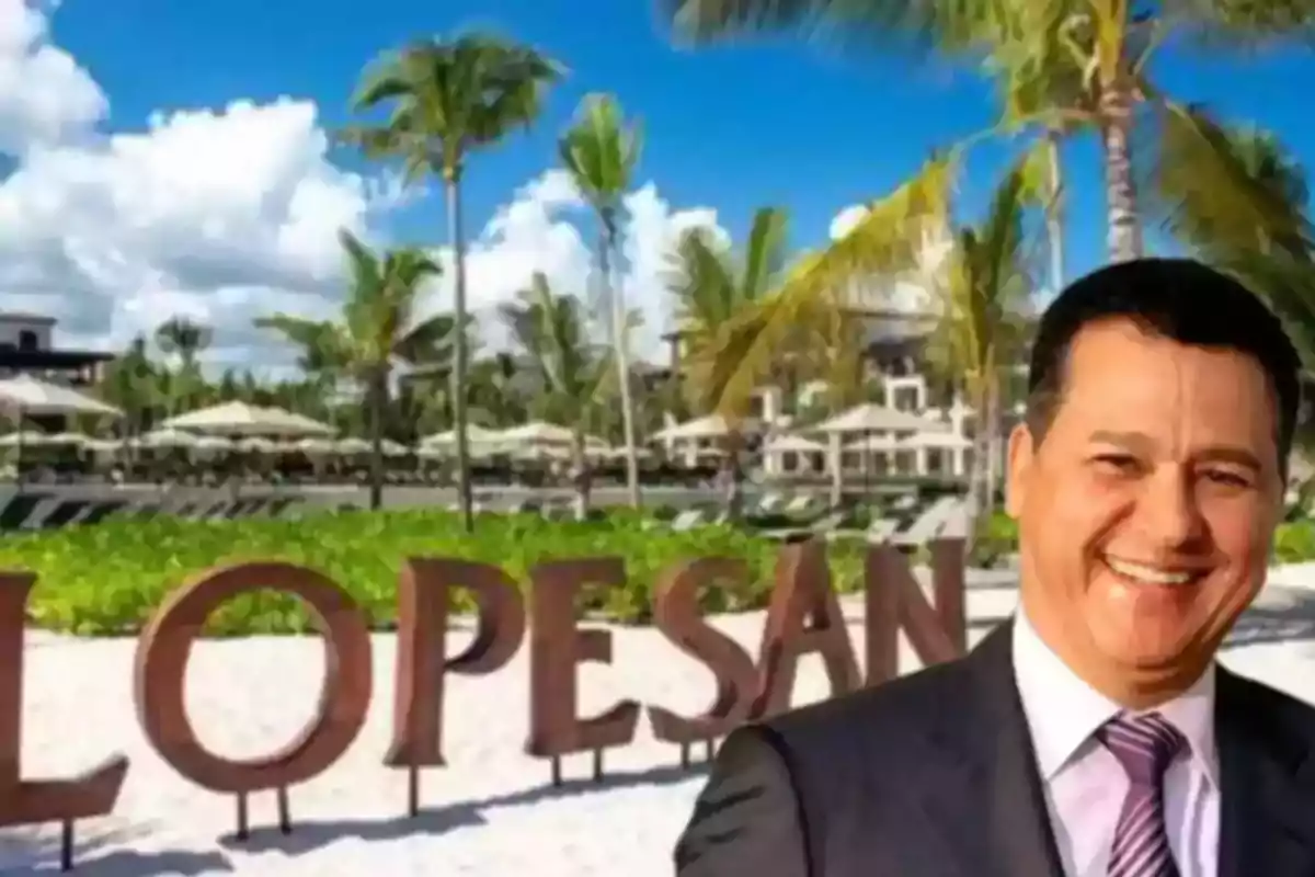 Hombre sonriente con traje frente a un letrero de Lopesan en una playa con palmeras y sombrillas Hombre sonriente con traje frente a un letrero de Lopesan en una playa con palmeras y sombrillas