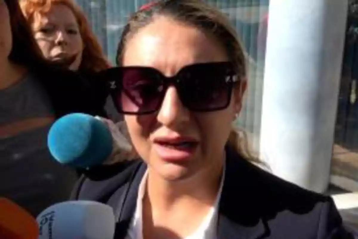 Mujer con gafas de sol grandes hablando frente a varios micrófonos en un espacio exterior Mujer con gafas de sol grandes hablando frente a varios micrófonos en un espacio exterior