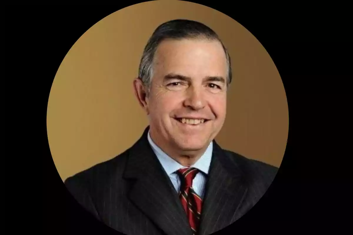 Hombre de mediana edad con cabello corto y canoso, vestido con traje oscuro, camisa azul y corbata a rayas, sonriendo frente a un fondo marrón. Hombre de mediana edad con cabello corto y canoso, vestido con traje oscuro, camisa azul y corbata a rayas, sonriendo frente a un fondo marrón.