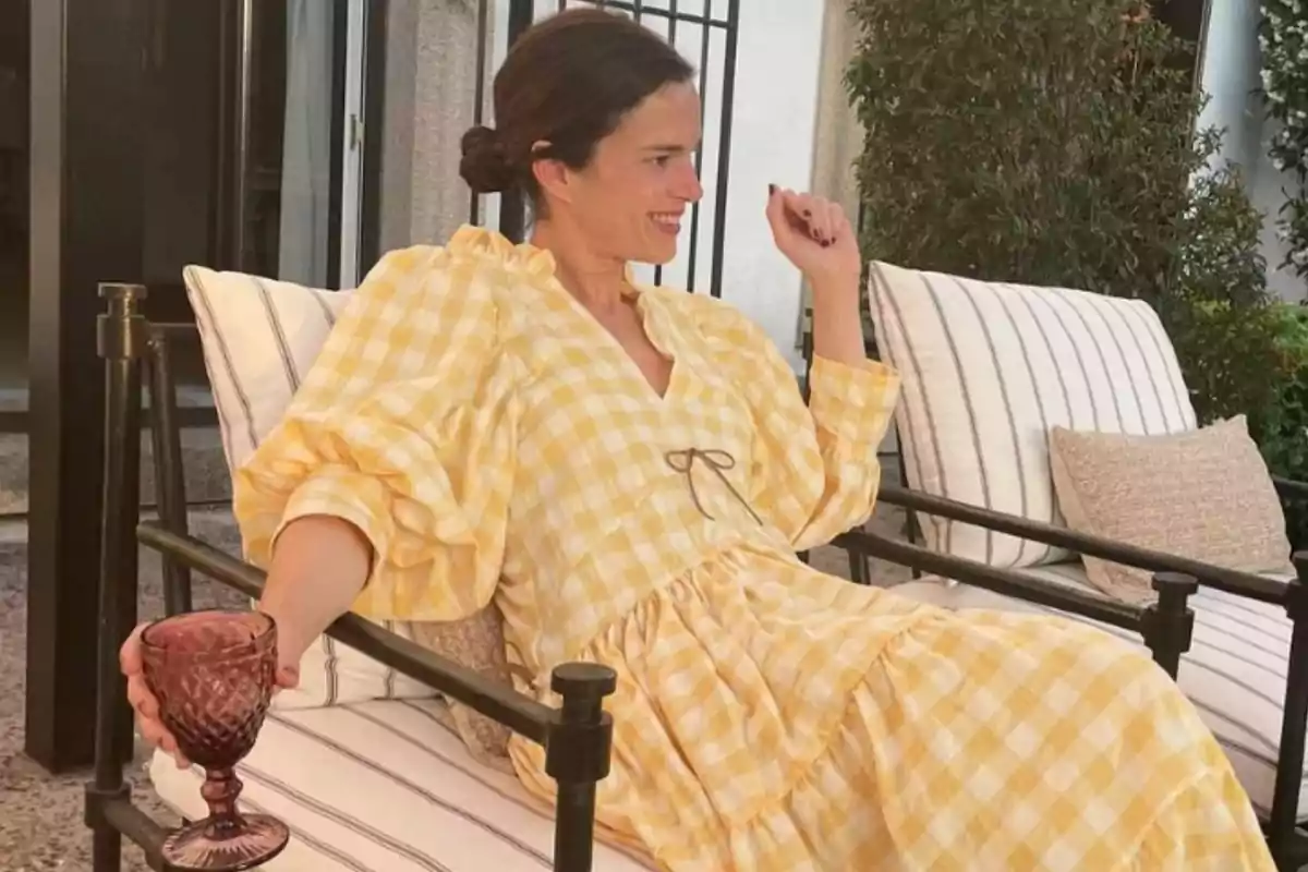 Mujer sentada en un sillón al aire libre con un vestido de cuadros amarillos y blancos sosteniendo una copa morada y sonriendo Mujer sentada en un sillón al aire libre con un vestido de cuadros amarillos y blancos sosteniendo una copa morada y sonriendo