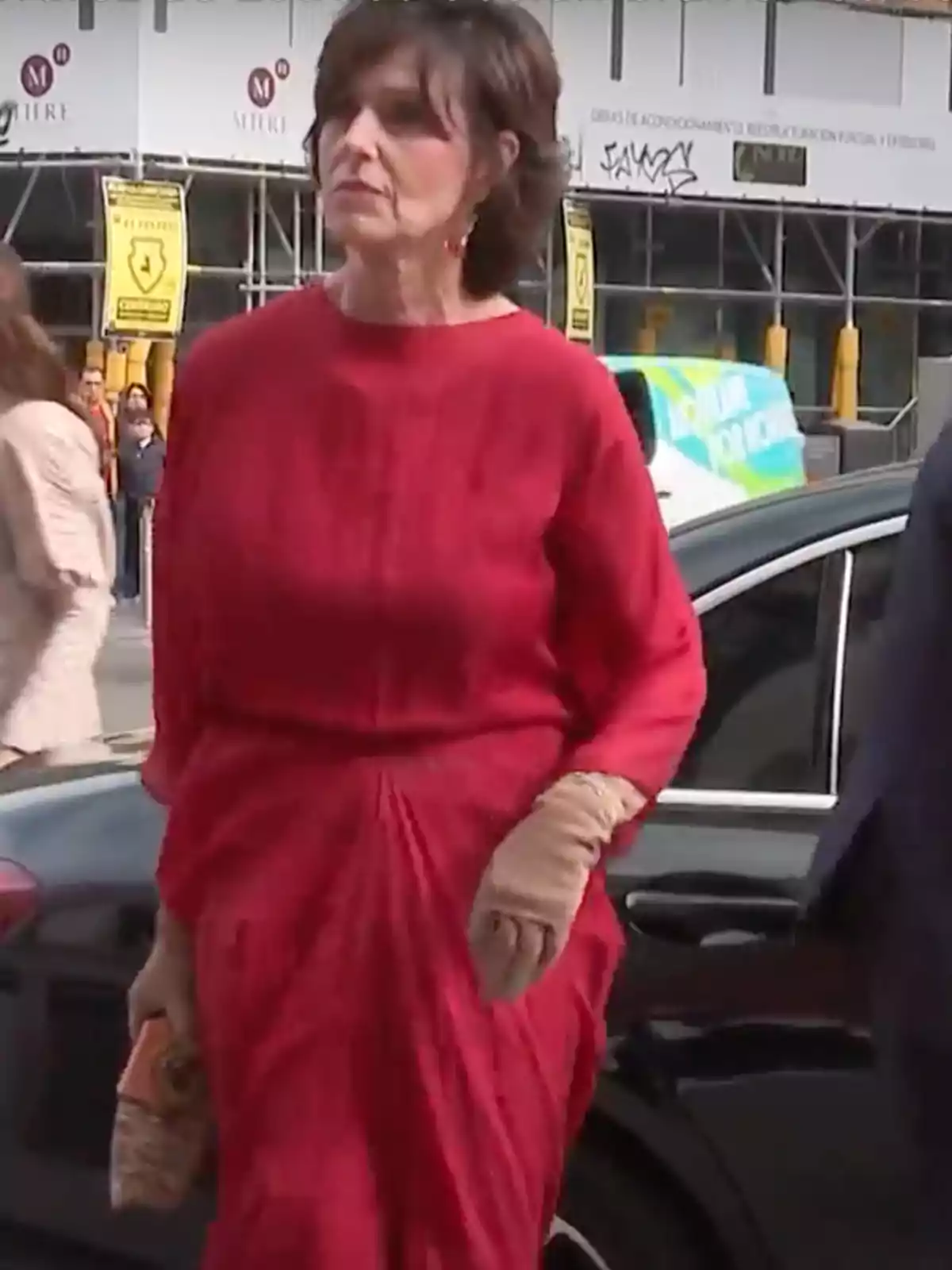 Mujer de cabello corto vestida con un vestido rojo y guantes beige caminando junto a un automóvil en una calle urbana Mujer de cabello corto vestida con un vestido rojo y guantes beige caminando junto a un automóvil en una calle urbana