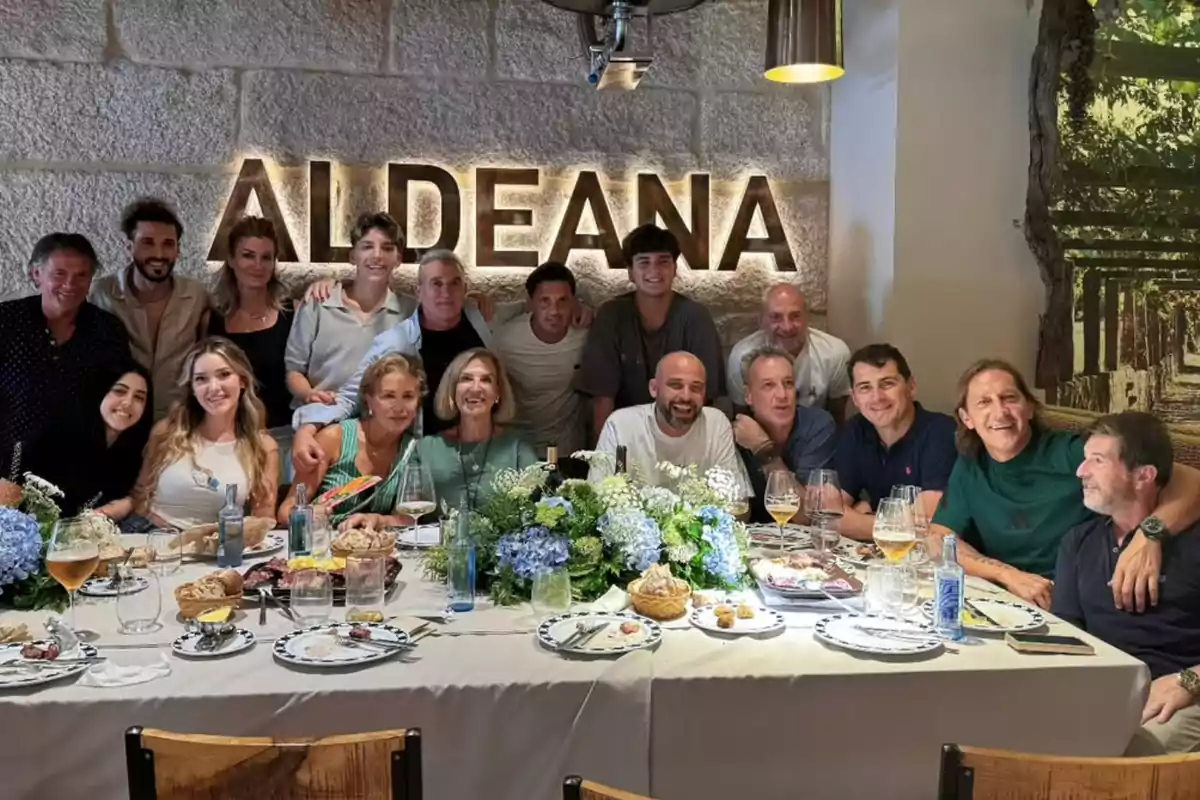 Un grupo de personas sonríe alrededor de una mesa decorada con flores y comida en un restaurante con la palabra ALDEANA iluminada en la pared de fondo