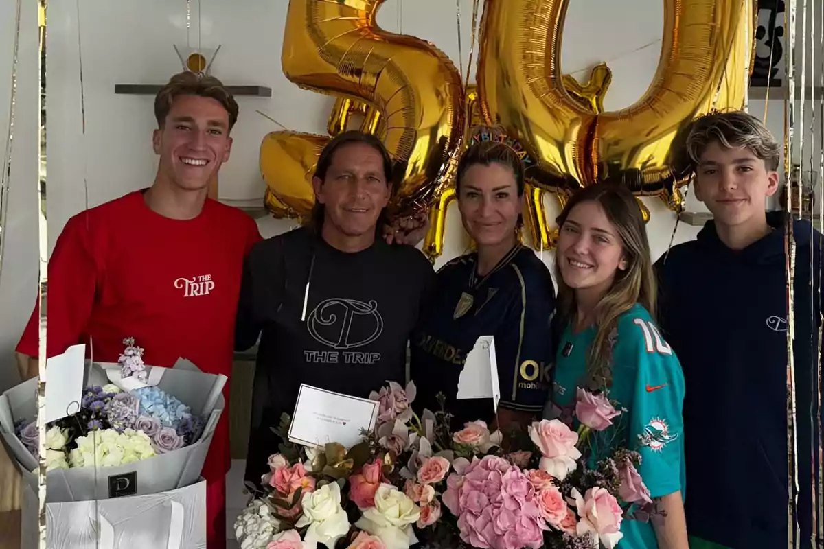 Cinco personas sonríen frente a globos dorados con el número 50 y arreglos de flores, celebrando un cumpleaños. Cinco personas sonríen frente a globos dorados con el número 50 y arreglos de flores, celebrando un cumpleaños.