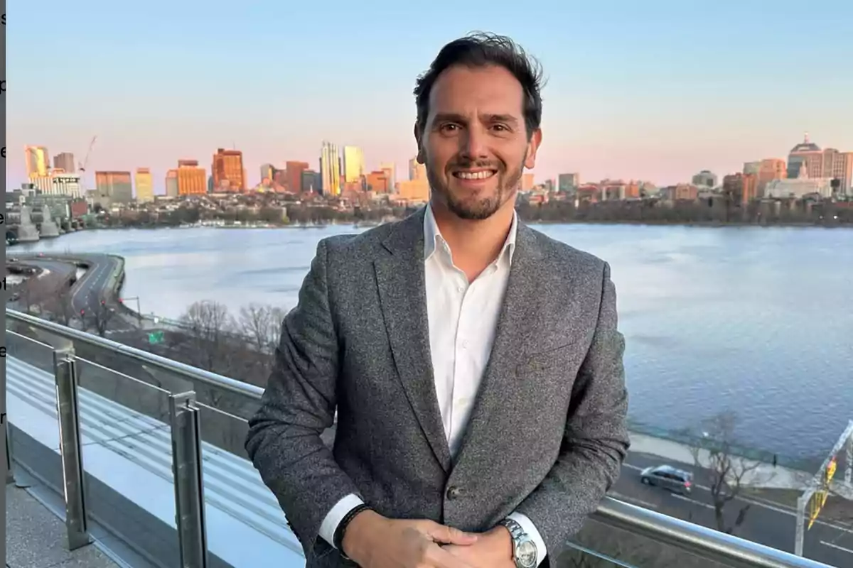 Hombre con saco gris y camisa blanca sonriendo en una terraza con vista a un río y una ciudad al atardecer Hombre con saco gris y camisa blanca sonriendo en una terraza con vista a un río y una ciudad al atardecer