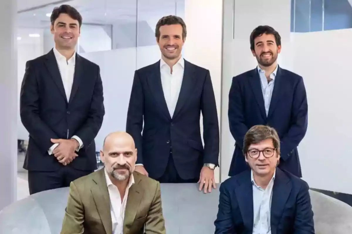 Cinco hombres vestidos con trajes formales posan en una oficina moderna, tres de ellos están de pie y dos sentados en un sofá. Cinco hombres vestidos con trajes formales posan en una oficina moderna, tres de ellos están de pie y dos sentados en un sofá.
